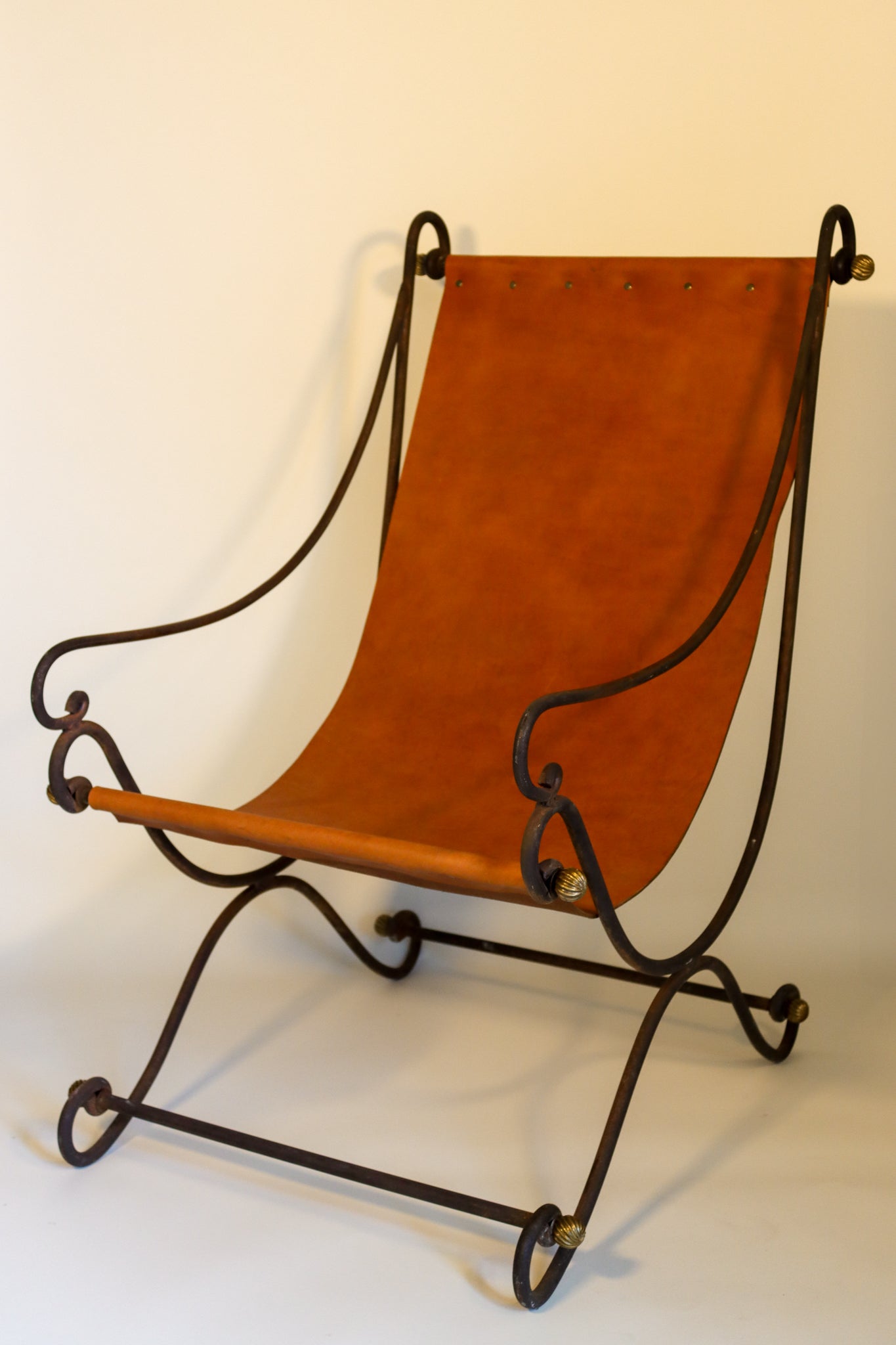 A sling arm chair with a black metal frame with gilt-metal roundels, formed with scrolling supports and a curule base, with burnt-orange leather sling seat and back.